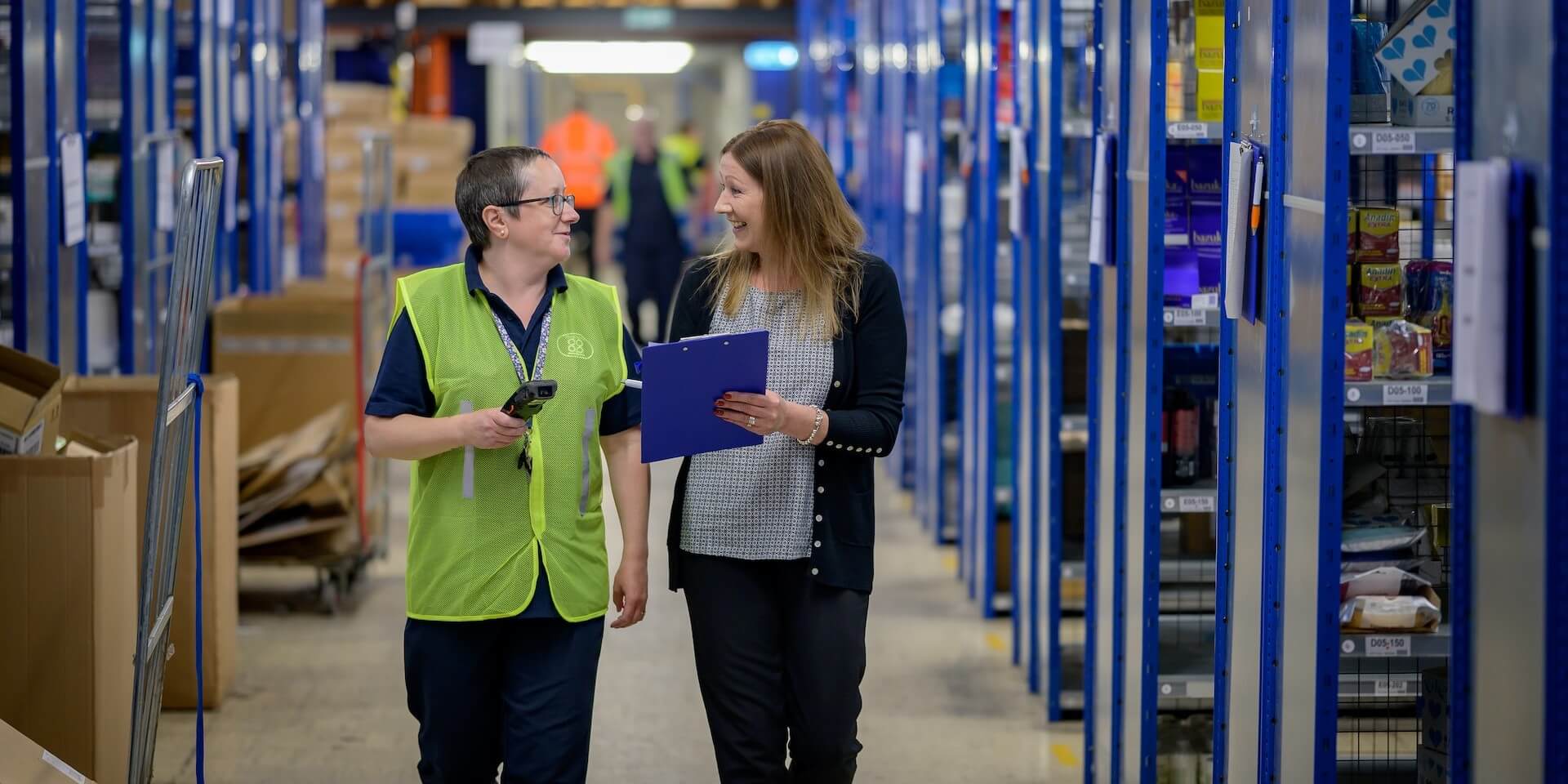 two women walk down a long hallway with clipboard